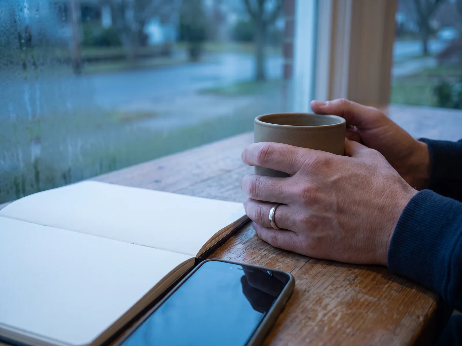 Two hands around a mug by a rainy window, with an open notebook and a phone on the table.