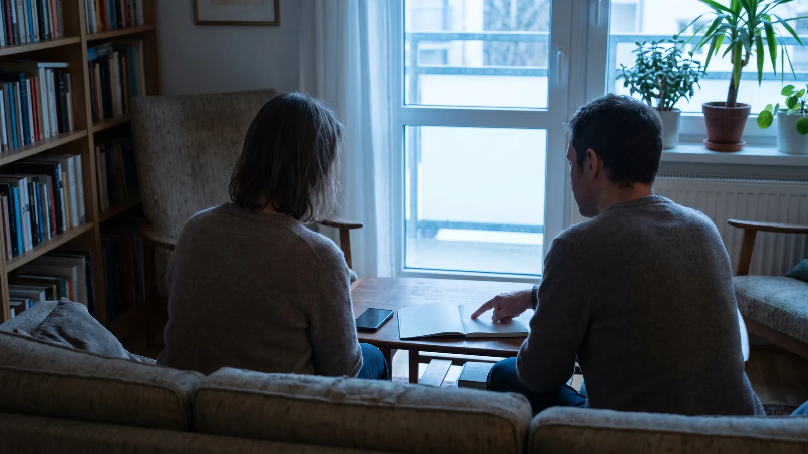 Two people on a sofa, seen from behind, reading an open notebook together on a coffee table by a window.