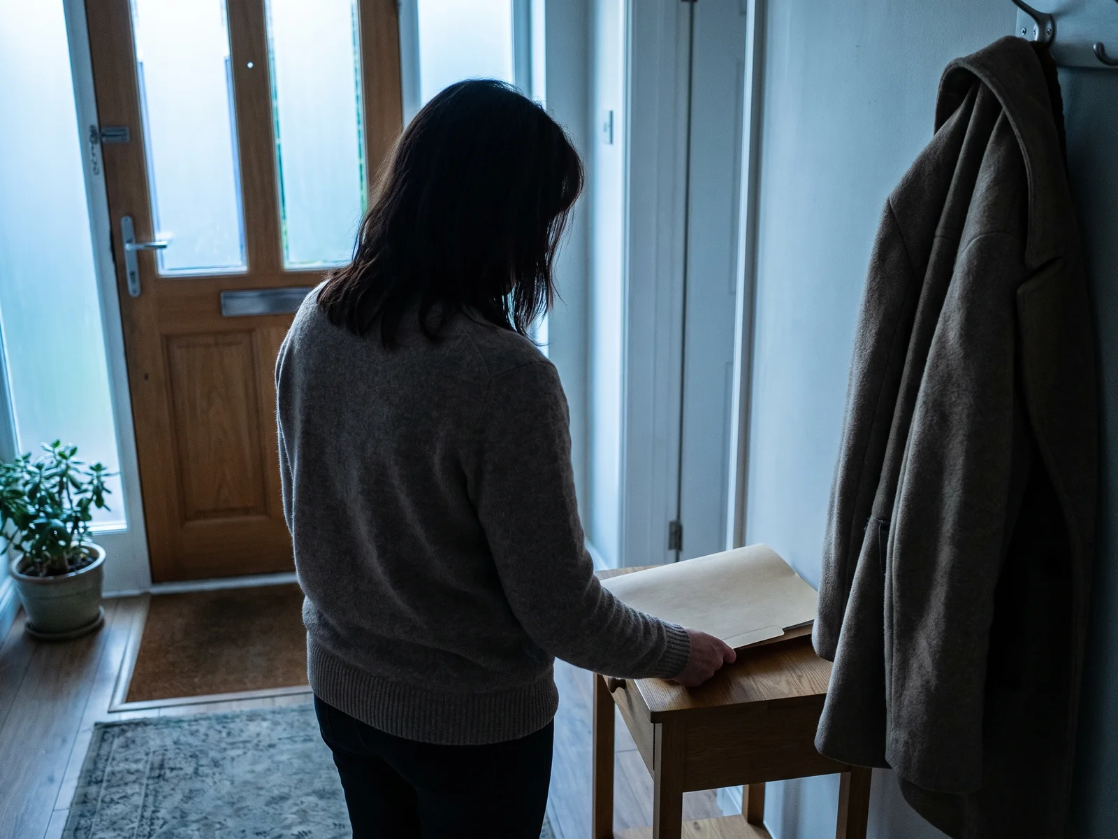 A homeowner, seen from behind, sets a folder of claim papers on a small entry table by the front door, a coat on the hook beside her.